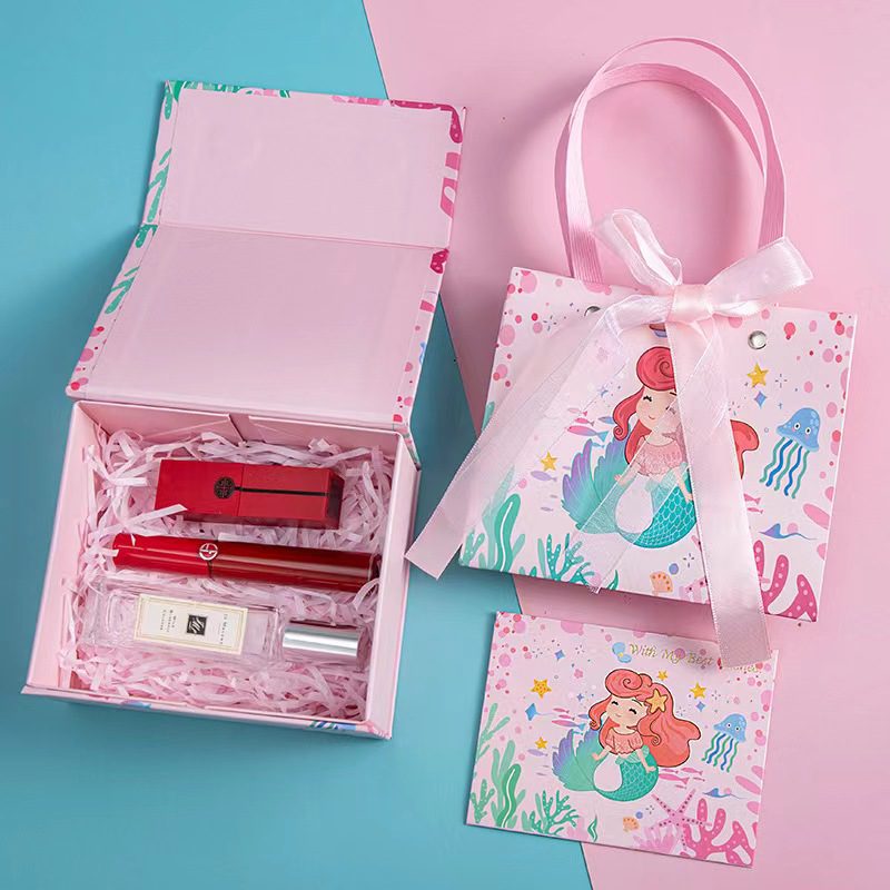 This image features a beautifully designed gift box set with a mermaid theme. The set includes an open gift box on the left, revealing neatly arranged contents such as a red lipstick, a red perfume bottle, and a clear bottle of perfume, all cushioned with pink shredded paper. To the right, there is a matching pink gift bag with handles and a bow, adorned with an illustration of a mermaid. Below the gift bag, there is a matching greeting card with the same mermaid design. The background is split into two colors, blue on the left and pink on the right, complementing the color scheme of the gift set