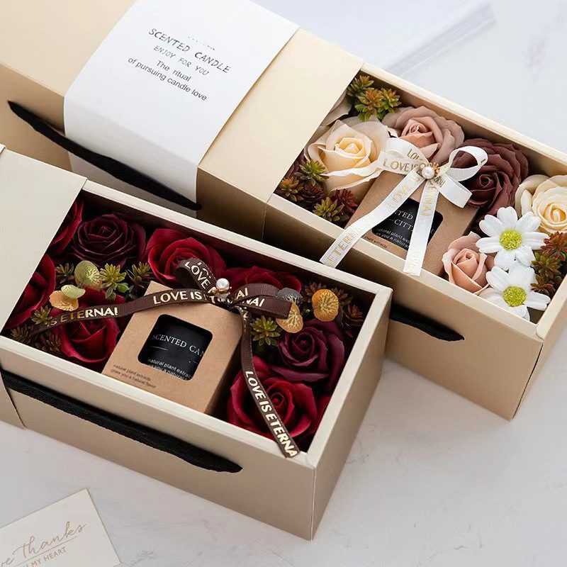 This image features two elegantly packaged gift boxes perfect for Mother's Day gift ideas for women. Each box is filled with a variety of artificial flowers and a scented candle, providing a luxurious and thoughtful presentation. The left box showcases deep red roses surrounding a black scented candle, adorned with a ribbon that reads 'Love is Eternal.' The right box displays a mix of beige, brown, and white flowers, including roses and daisies, encircling a similarly packaged scented candle with a white ribbon. Both boxes have a sophisticated beige exterior with a minimalist design and black handles, making them an ideal choice when considering what is a some gift idea for women