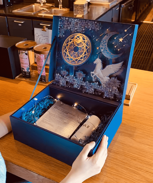 This image shows an intricately designed gift box with a celestial theme. The box is open, revealing an elaborate interior decorated with a night sky scene featuring a golden geometric circle, a crescent moon, stars, and a white Pegasus. Inside the box, nestled in blue shredded paper, are a bottle of SK-II facial treatment essence and a set of skincare masks. The box is further adorned with fairy lights, adding a magical touch to the presentation. The scene is set on a wooden table in a cozy, well-lit room, with the box being held by a person's hands, ready to be given as a luxurious gift.