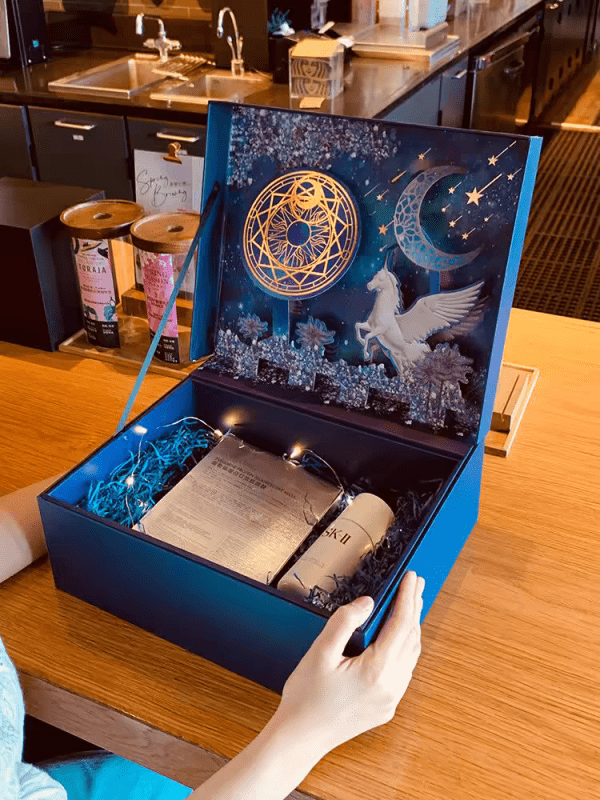 This image shows an intricately designed gift box with a celestial theme. The box is open, revealing an elaborate interior decorated with a night sky scene featuring a golden geometric circle, a crescent moon, stars, and a white Pegasus. Inside the box, nestled in blue shredded paper, are a bottle of SK-II facial treatment essence and a set of skincare masks. The box is further adorned with fairy lights, adding a magical touch to the presentation. The scene is set on a wooden table in a cozy, well-lit room, with the box being held by a person's hands, ready to be given as a luxurious gift.